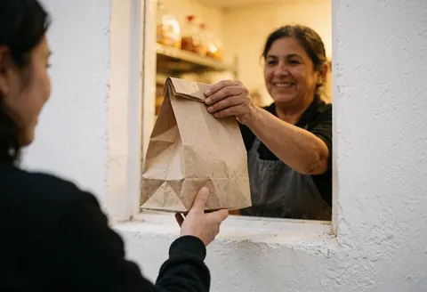 A smiling bakery owner hands a food bag to a customer through a window with warm lighting and a white wall.
