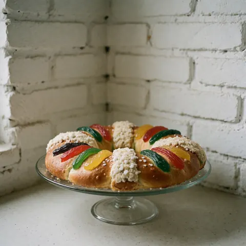 A traditional Rosca de Reyes sweet bread topped with colorful candied fruits and powdered sugar, displayed on a glass counter against a brick wall.