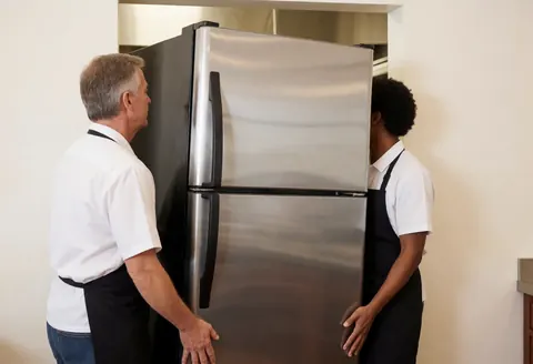 Workers installing a large stainless steel refrigerator in a modern commercial kitchen setting