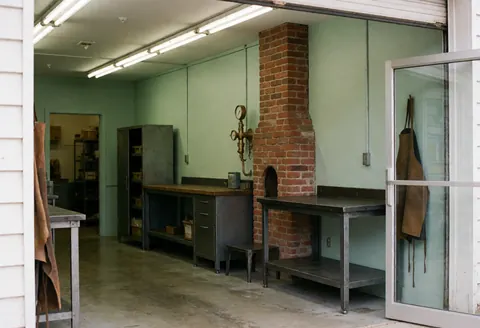 Front view of a brick workshop building with a chimney, entrance door, and windows, representing a small manufacturing facility.