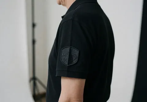 Close-up of a business professional in a black polo shirt showing an embroidered patch on the sleeve against a white studio background.