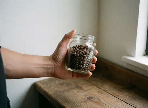 Close-up of fresh roasted coffee beans inside a glass jar placed on a wooden surface with natural light coming from a nearby window