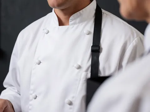 Detailed view of the textured fabric, stitching, and buttons on a professional chef's jacket in a commercial kitchen setting