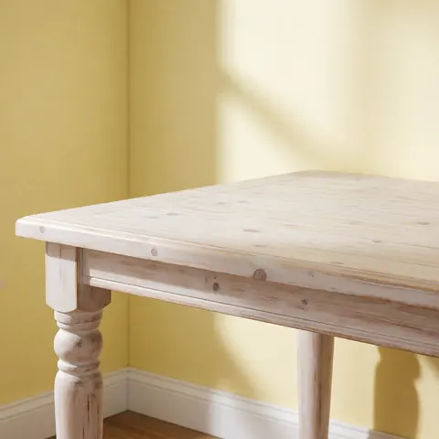 A white painted pine farmhouse dining table featuring turned legs and visible wood grain, set against a pale yellow wall in a rustic interior setting.