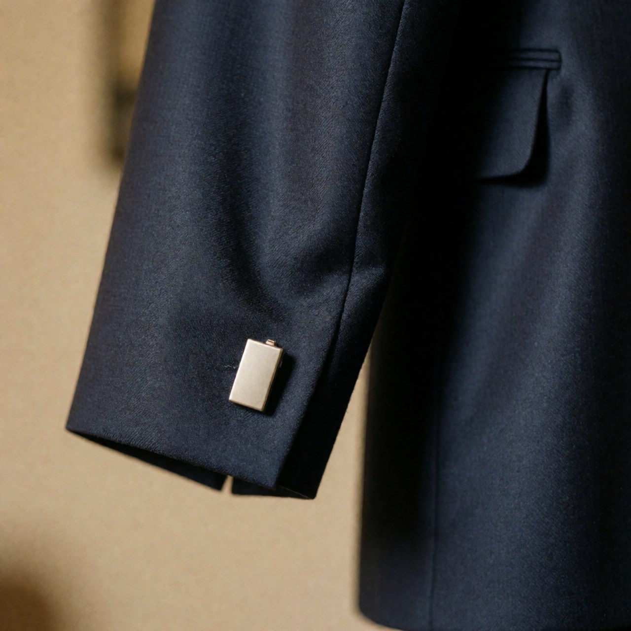 Detailed view of a navy blue suit jacket showing the collar, sleeve button, and fine fabric texture with warm lighting and a blurred background.