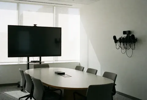 Empty modern conference room featuring an oval table, large screen, and professional microphones with natural light and white walls.