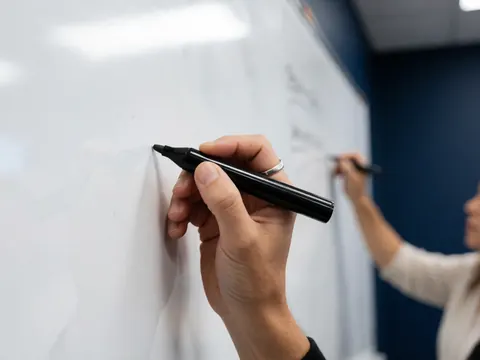 Close-up of a hand with a ring writing on a whiteboard with a black marker while another person writes in the background against a dark blue wall