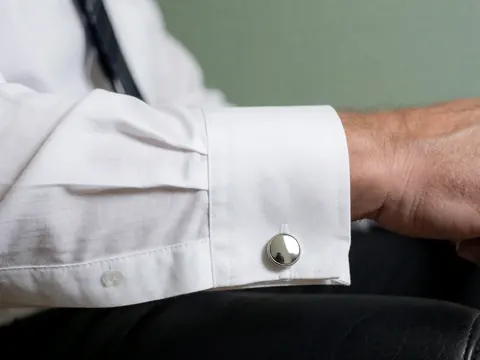 Macro shot of a white dress shirt sleeve with silver cufflinks resting on black leather against a pale green background