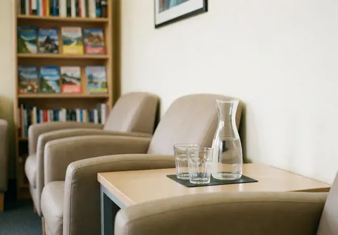 A clean and bright waiting room interior featuring wooden shelves, books, and a table with water carafes for language school students
