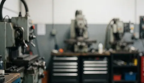 A blurred gray industrial workshop background featuring out-of-focus machinery, tools, and metal surfaces suitable for text overlay in manufacturing contexts