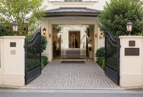 The main entrance of a hotel featuring an elegant iron gate, paved walkway, and surrounding greenery leading to the glass doors.