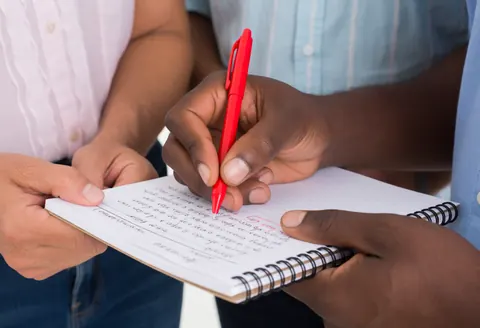 Teacher using a red pen to correct a Spanish exercise in a notebook while a student watches attentively during a language class session.