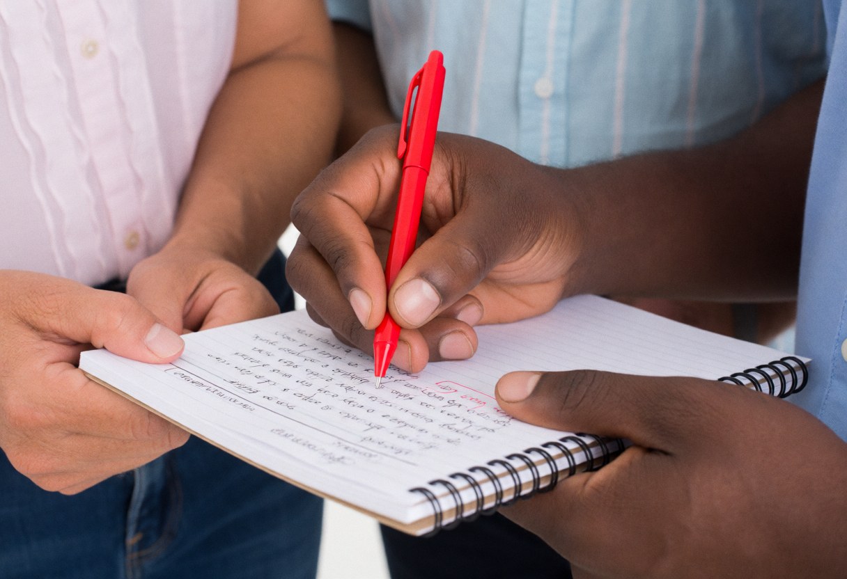 Teacher using a red pen to correct a Spanish exercise in a notebook while a student watches attentively during a language class session.