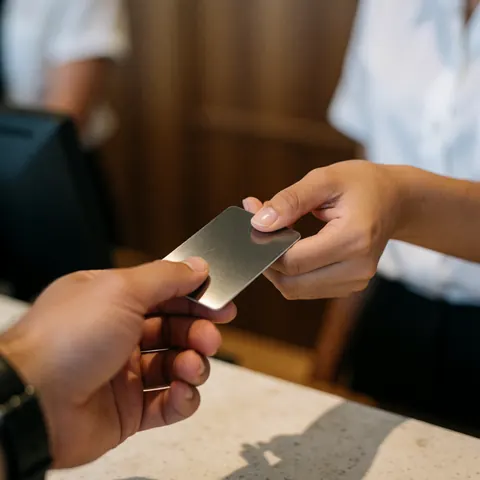 A close-up view of a hand handing a metal hotel room key to another hand at a wooden reception desk, symbolizing check-in service and hospitality.