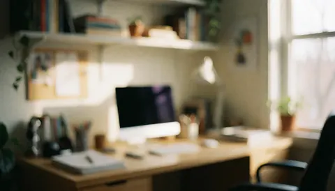 A softly blurred background of a home office desk featuring a computer, notebook, pen, and shelves with plants, illuminated by warm natural light from a window, ideal for insurance text overlays.