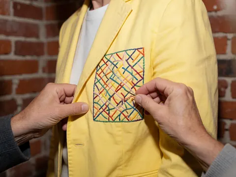 Close-up of hands performing colorful embroidery on a yellow jacket against a brick background, highlighting artisanal textile details for corporate branding.