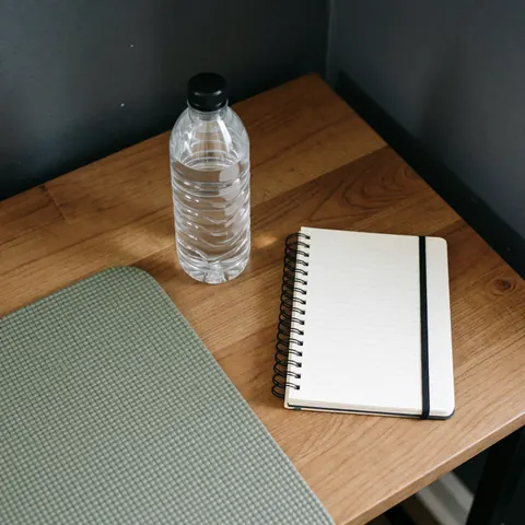 Wellness kit featuring a water bottle, spiral notebook, and yoga mat arranged on a wooden table against a gray wall, promoting a healthy and productive lifestyle.