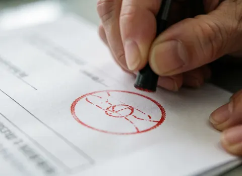 Close-up of an adult hand applying a circular red stamp to an office document with a blurred background and clinical lighting.