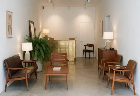 A spacious and calm hotel lobby featuring modern wooden furniture, bronze accents, white walls, and ambient lighting, creating an inviting atmosphere for guests.