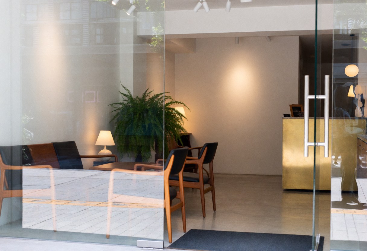 Modern boutique hotel entrance featuring a bronze reception desk, teak furniture, white walls, and a large indoor fern plant.