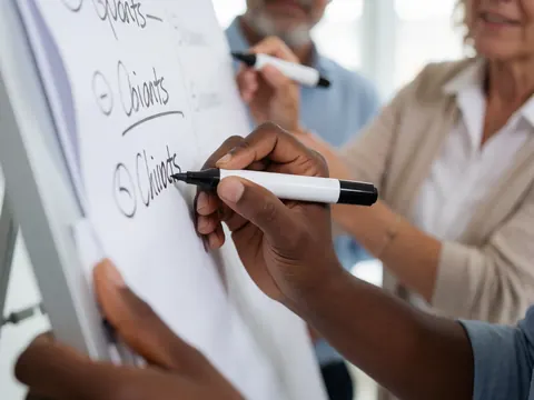 Close-up of hands writing on a whiteboard with a marker during a professional meeting with colleagues in the background