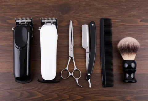 Top view of barber scissors, razor, comb, brush, and clippers on a dark wooden surface with soft professional lighting