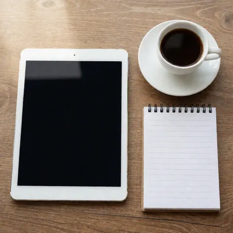 Top view of a tablet, white coffee cup, and notebook on a wooden desk, ideal for human resources and productivity.