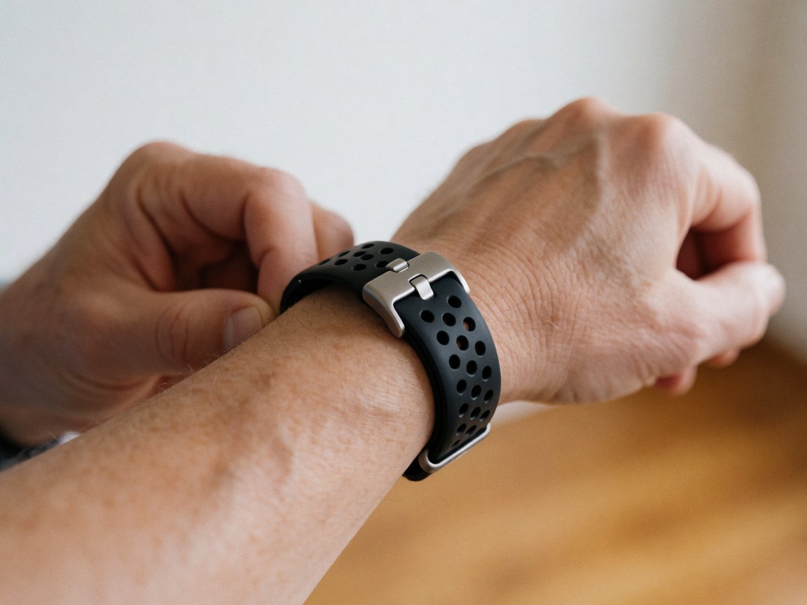 Detailed view of a smartwatch clasp featuring a black perforated strap and shiny metal, held in hand against a warm wooden background with natural light.