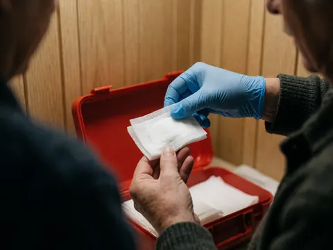 A gloved hand pulls a white sterile bandage from a red first aid kit against a light wood wall background with natural lighting.