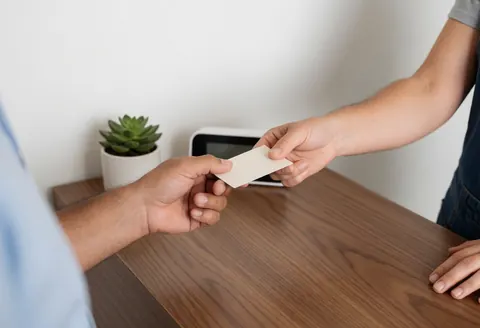 Two people exchange business cards on a wooden desk with a plant and digital clock in the background