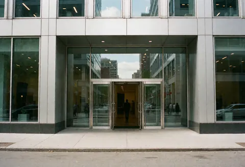 Modern corporate building entrance featuring glass doors, city reflections, and a person entering. Clean professional architecture suitable for business and agreements.