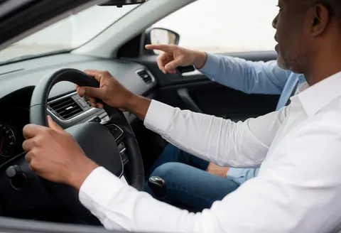 Interior view of a car showing a driving instructor pointing ahead while a student keeps hands on the steering wheel during a daytime lesson