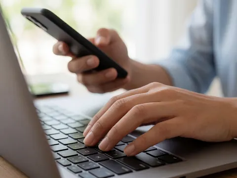 Close-up of hands typing on a laptop keyboard while holding a smartphone in a professional workspace with natural light and selective focus on the fingers.
