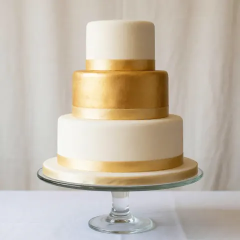 A white wedding cake featuring elegant gold accents and intricate detailing, presented on a clear glass stand against a neutral background for product display.