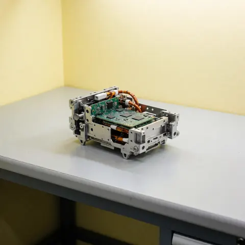 Metal electronic subassembly with circuits and wires on a gray workbench against a light yellow wall with neutral professional lighting.