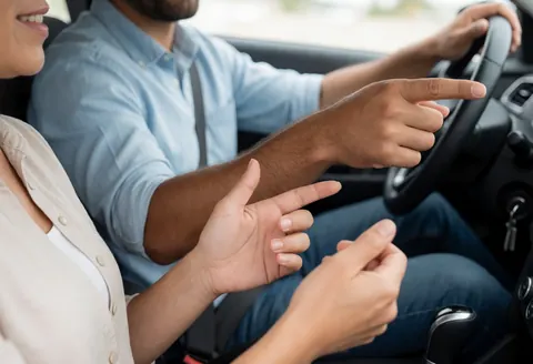 A driving instructor sits in the passenger seat of a car, pointing ahead to guide the student driver who is holding the steering wheel during a daytime lesson.