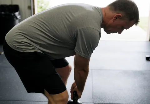 A man lifts a kettlebell with focus in a gym with rubber flooring and natural light, showing realistic and professional fitness action.