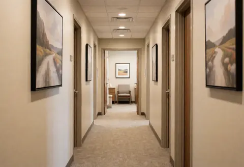 A quiet spa corridor featuring framed wall art, beige waiting chairs, and treatment room doors with warm lighting and wooden accents.