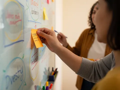 Hand placing an orange sticky note with inclusion text on a whiteboard with diagrams while another person writes in the background during a professional training session
