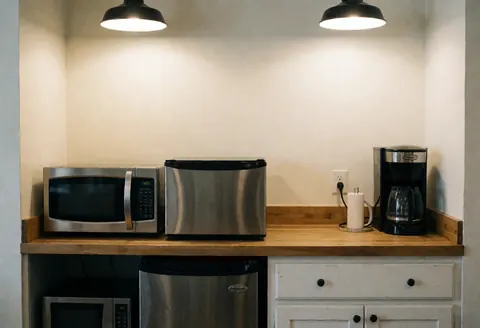Office kitchen featuring microwave, refrigerator, and coffee maker on a wooden table with warm walls and ceiling lights.