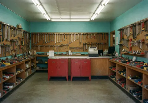 Hardware store interior featuring turquoise walls, concrete floor, wooden shelves stocked with tools, red boxes, and a vintage cash register.
