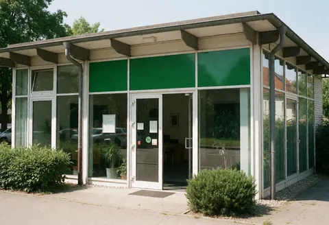 Modern solar energy office building featuring a glass facade, glass entrance door, green signage, and landscaped bushes on a sunny day.