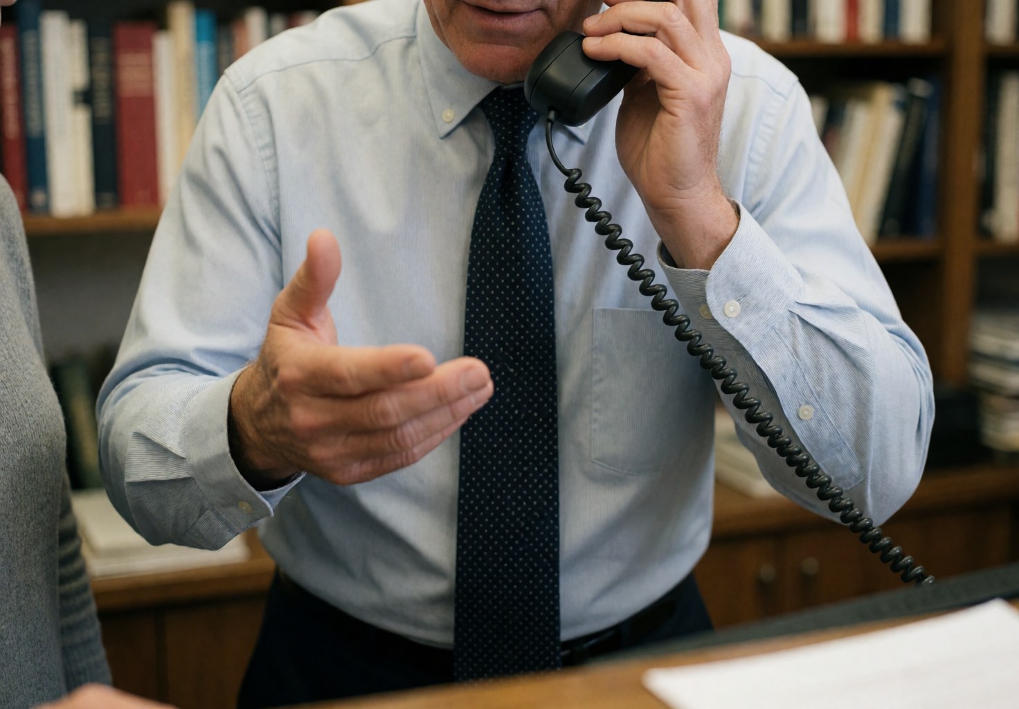 A male receptionist in an office setting holding a phone to his ear and gesturing with his hand while speaking, with bookshelves visible in the background.
