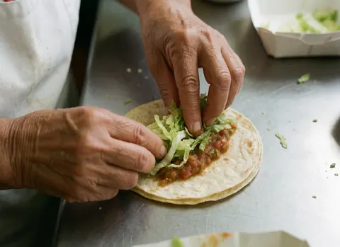 Hands assembling a fresh taco with lettuce and salsa on a shiny metal kitchen counter, showcasing crisp textures and vibrant ingredients for fast food preparation.