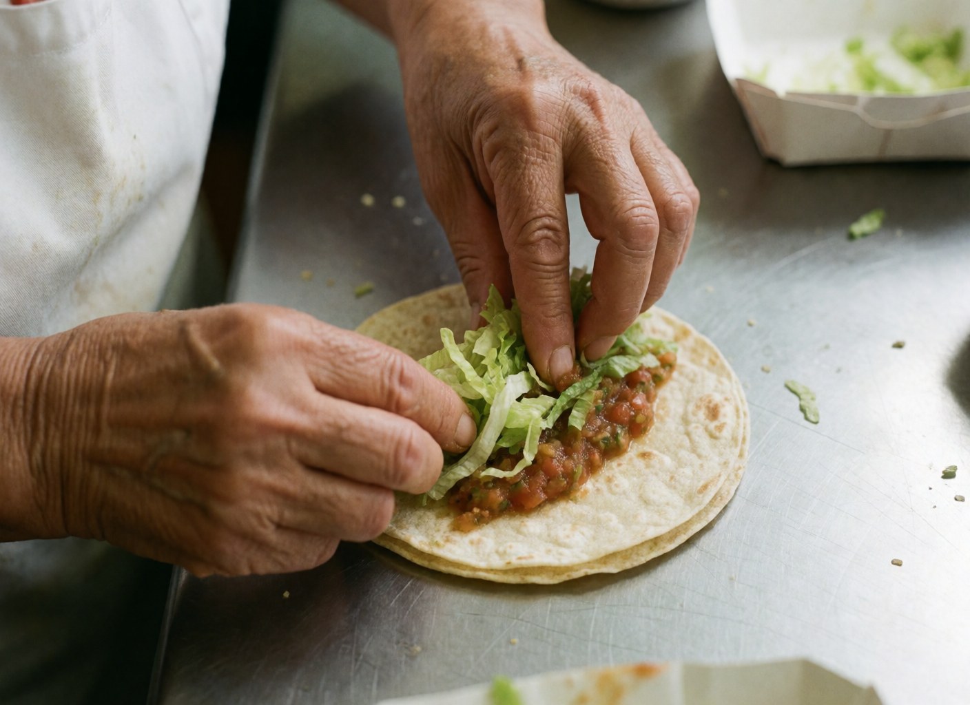 Hands assembling a fresh taco with lettuce and salsa on a shiny metal kitchen counter, showcasing crisp textures and vibrant ingredients for fast food preparation.
