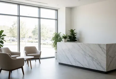 Modern office reception featuring a white marble desk, comfortable chairs, green plants, and large windows with natural light in a clean minimalist space.