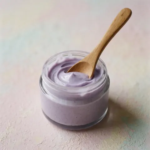Glass jar of lavender cream with a wooden spoon on a soft pastel background with natural lighting