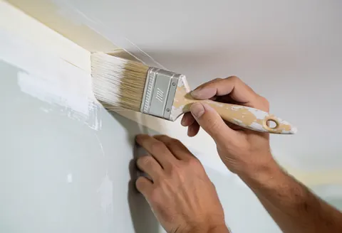 Close-up of a hand using a paintbrush to apply bright, clean paint to a wall near masking tape, showing ceiling and wall details under soft professional lighting