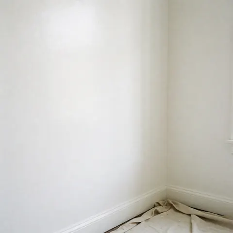 Top view of a white interior wall with baseboard, paint cloth on the floor, and a glossy finish for real estate and home decor.