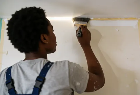 A professional painter applies white paint to an interior wall with a brush in a real work environment with natural light and partially painted walls.